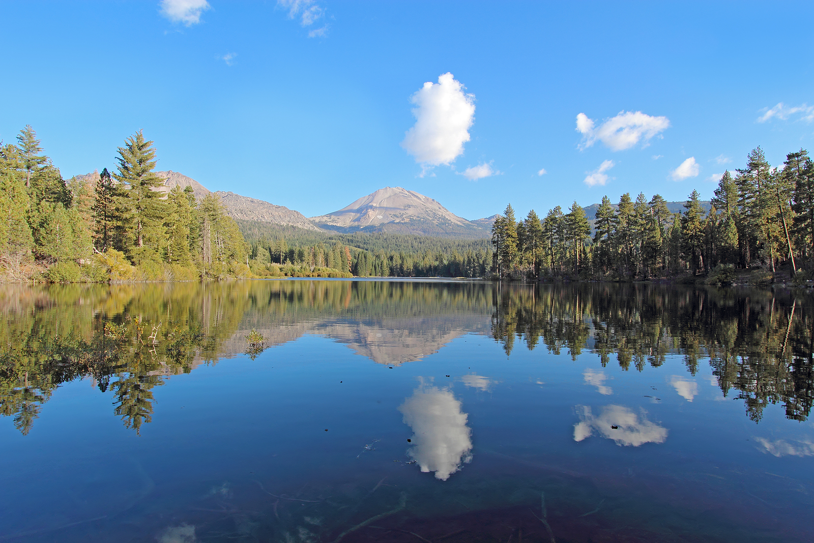 Wide-angle view of Lassen Peak trees and white clouds reflected in the calm waters on Manzanita Lake near the entrance to Lassen Volcanic National Park in northern California